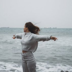 A woman stands with arms spread by the sea, embracing the freedom of a windy, overcast day.