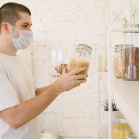 A young man with a facemask in a modern kitchen reviewing pantry items.